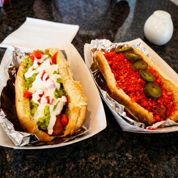 Two gourmet hot dogs, one with guacamole and tomatoes, the other with red spicy crumbs and jalapeños.