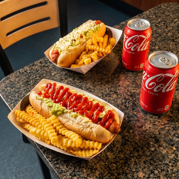 Two hot dogs with fries and two Coca-Cola cans on a speckled countertop.