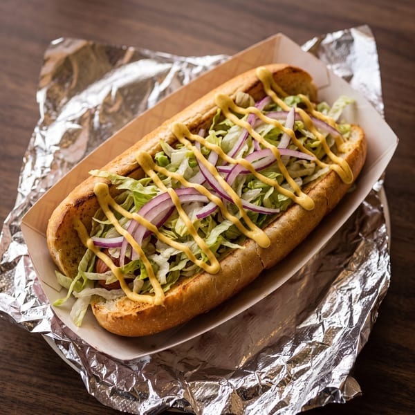 Hot dog in a bun with lettuce, red onions, and mustard, served in a paper tray on foil.