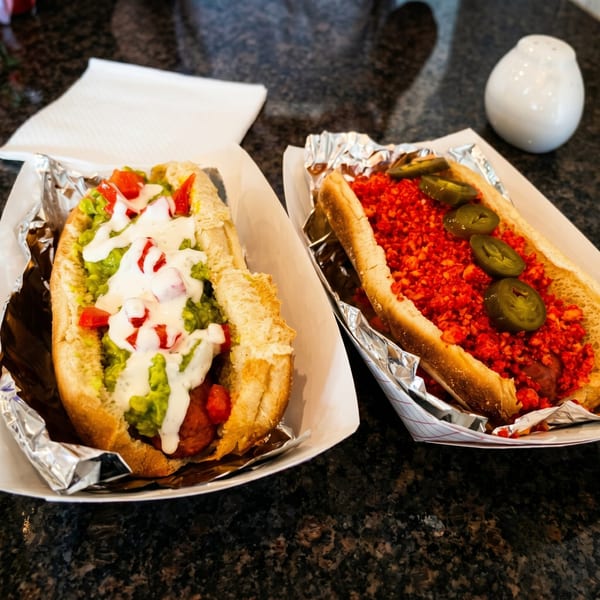 Two gourmet hot dogs in foil-lined trays, one with guacamole and the other with red spicy toppings.