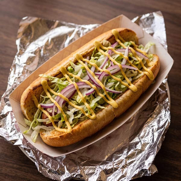 Hot dog in a bun with lettuce, red onions, and mustard, served in a paper tray on foil.
